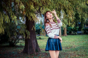 身穿條紋毛衣和藍色裙子的年輕女子站在公園的樹下。 — 樹 壁紙