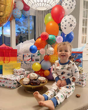 一個微笑的幼兒穿著Paw Patrol睡衣，被生日氣球、禮物和冬甩圍繞。