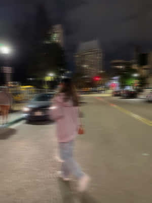 一個穿著粉紅色連帽衫的人，在夜晚的城市街道上行走，帶有模糊的動作和發光的車燈。