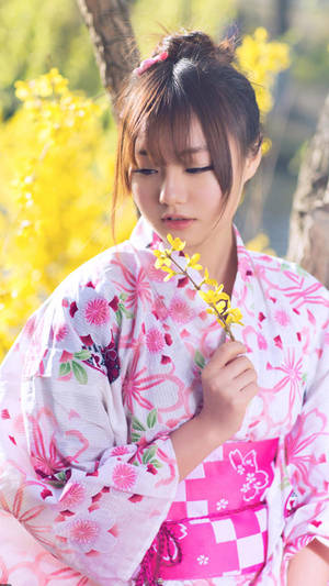 年輕女子穿著粉紅色花卉和服，在春天花園中手持黃色花枝。 — 日本 壁紙