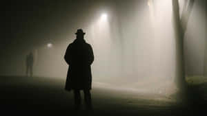 穿著 trench coat 和 fedora 帽的男子在夜間霧氣瀰漫的公園中獨自站在路燈下。 — 恐怖電影 壁紙