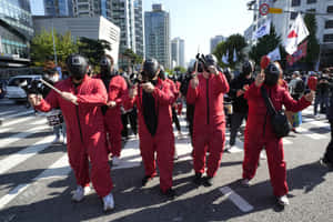 穿著紅色連身衣和黑色面具的抗議者手持樂器在城市街道上游行。 — MacBook Pro M3 壁紙