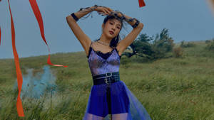 一位身穿藍色閃亮服裝的女性在田野中擺姿勢，周圍有紅色絲帶和藍色煙霧。
