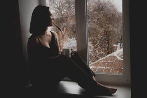 剪影中的女人在窗邊拿著杯子，看著被雪覆蓋的樹木