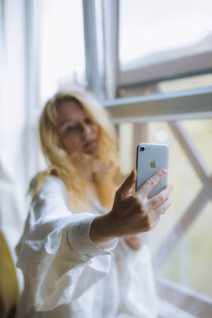 金髮女子穿白色襯衫在窗邊用銀色iPhone自拍。