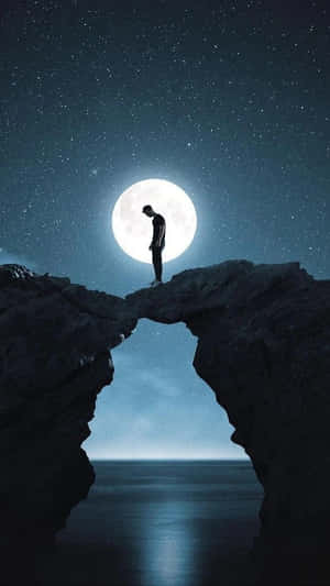 夜晚，一個人站在岩石拱門上，滿月高懸於海面上空的剪影。 — 月亮 壁紙