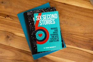 藍綠色書本名為 Six Second Stories 放在木桌上，下方有藍色筆記本。 — 書本美學 GIF 壁紙