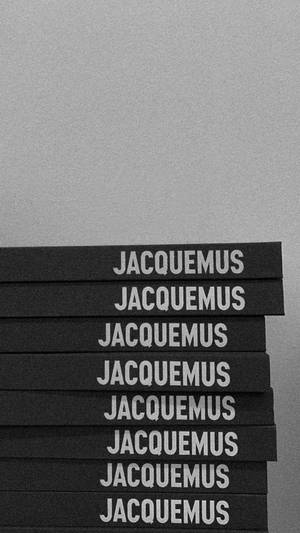 一堆黑色書籍，上面有白色 Jacqumus 文字，背景為灰色。
