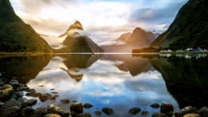 參差不齊的綠色山脈倒映在日出時分的平靜峽灣中，前景岩石嶙峋。