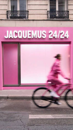 一位身穿粉紅色的單車手在城巿街道上經過一間粉紅色的 Jacquemus 商店。