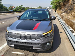 灰色Jeep Compass，帶紅色條紋，停在陽光明媚的高速公路旁，靠近綠樹和山丘。