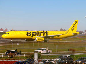 黃色Spirit Airlines飛機在機場停機坪上，背景有城市天際線和前景的汽車。
