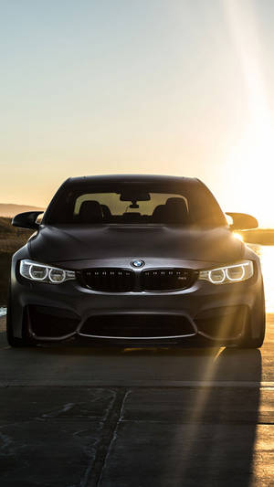 啞光黑色BMW M4在日落時分，車頭燈發光，行駛在鋪砌的道路上。