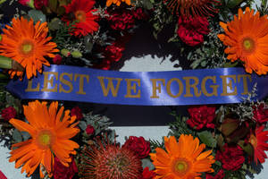 一個花圈，有橙色和紅色的花朵，以及一條寫有「LEST WE FORGET」的藍色絲帶。