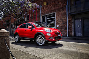 紅色Fiat 500X SUV停泊在陽光明媚的城市街道上，靠近磚砌建築和開花的樹。