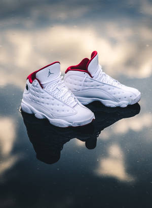 白色Jordan 13運動鞋帶紅色點綴，在潮濕路面上反射雲朵。