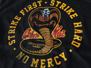 黑色T恤，上面有「Cobra Kai」眼鏡蛇標誌同「Strike First Strike Hard No Mercy」字樣。