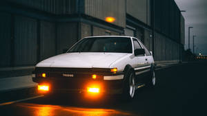 白色AE86跑車，帶有發光的頭燈，停泊在黃昏時潮濕的城市街道上。