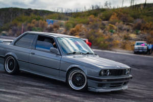 灰色BMW E30在賽道上飄移，後面有秋色山丘。