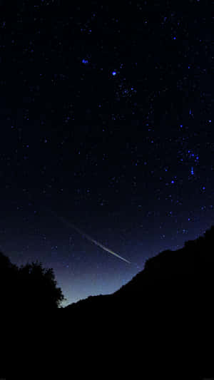 一顆流星在黑暗的山丘和樹木上方，劃過繁星點點的夜空。 — 夜空 壁紙