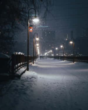 雪落在燈火通明的城市步道上，遠處有人行走。 — 夜晚 GIF 壁紙