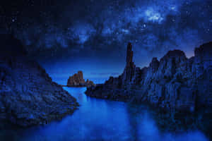 繁星點點的夜空，銀河在平靜的藍色海洋和岩石懸崖上空。