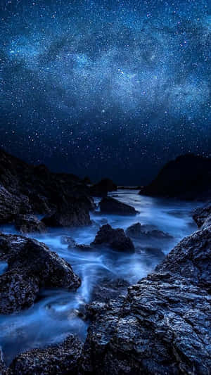 繁星點點的夜空，銀河在深色岩石之間流動的發光藍色海洋之上。 — 平板電腦 壁紙