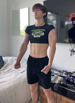 一個身材健美的年輕男子穿著遊戲主題短版上衣，站在有電視和遊戲設備的臥室裡。