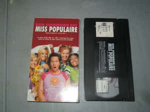 Rob Schneider的法國電影《Miss Populaire》的VHS錄影帶和封套，放在灰色表面上。 — 嘉麗姬莉 GIF 壁紙