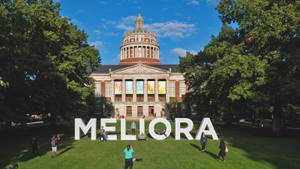 學生們聚集在草地校園廣場上，圍繞著拼出 MELIORA 的大型白色字母。 — 學校 壁紙