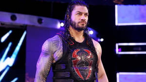 摔角手 Roman Reigns 身穿帶有紅色蜘蛛標誌的黑色背心，在紫色燈光下。