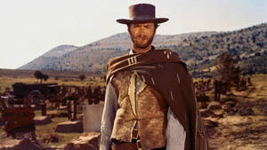 一個戴著帽子和披風的牛仔站在西部墓地中，背景有山。