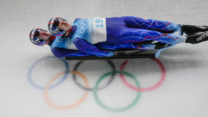 兩名雪橇運動員在冰上競賽，下方有彩繪的奧運五環。