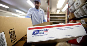 USPS分揀設施內的Priority Mail盒，背景有工作人員。