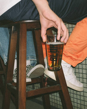 坐在木凳上手持啤酒杯的人，穿著牛仔褲和運動鞋。