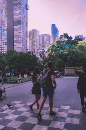 日落時分，人們在棋盤格廣場上行走，背景是城市建築和樹木。