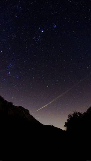 一顆明亮的流星在佈滿星星的夜空中劃出一道軌跡，下方是黑暗的山丘和樹木。 — 星星 壁紙