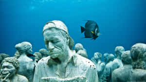 水下雕塑花園，有人物雕像和一條熱帶魚在附近游動。 — 水下 壁紙