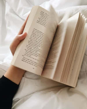 一隻手在白色床罩上拿著一本打開的書。 — 美學 壁紙