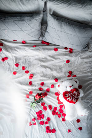 白色泰迪熊拿著心形，上面寫著「Be my Valentine」，在床上與玫瑰花瓣。