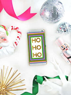 手機顯示 'HO HO HO'，被聖誕裝飾包圍，背景為白色毛絨。