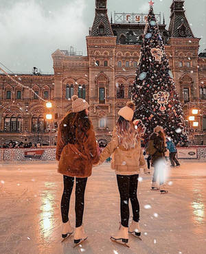 兩位身穿冬季外套的女性在聖誕樹附近的飄雪中溜冰。