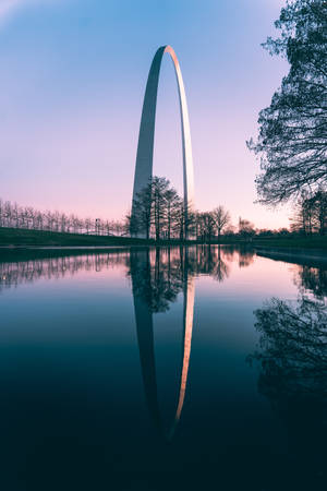 Gateway Arch 在暮色中倒映在水面上，周圍是光禿禿的樹木