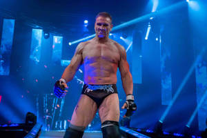 一位摔角手穿著黑色短褲和手套，在藍色舞台燈光下走著，火花四濺。