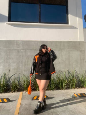 女性穿著黑色大學外套和太陽眼鏡，戶外站在混凝土牆附近。