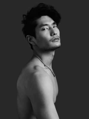 一個赤膊、深色頭髮、戴著項鍊的男人，在一張黑白工作室照片中向上看。