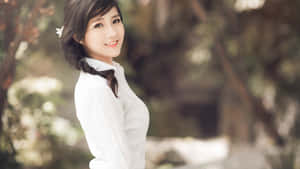 年輕女性身穿白色襯衫，在戶外微笑，背景是綠色樹葉。
