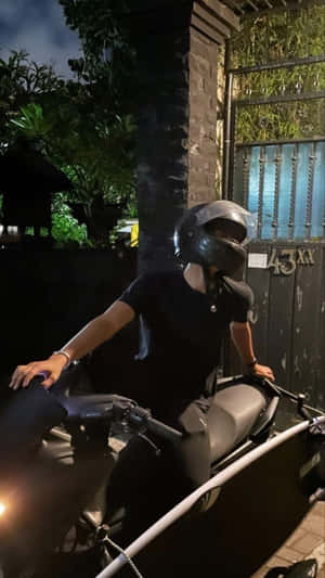 夜晚喺有閘門嘅入口附近，一個人喺黑色電單車上，旁邊有綠色植物。 — 夜晚 GIF 壁紙