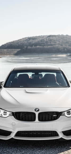 白色BMW M4停泊在湖邊，後面有樹木繁茂的山丘。 — iPhone 14 壁紙