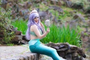 一個穿著美人魚服裝的人，紫色頭髮和藍綠色尾巴，坐在石牆上。